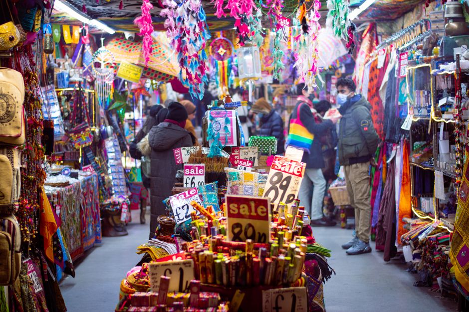 A busy street scene outside Camden Lock in London during daytime, featuring a large metal sign with the words 'Camden Lock' in bold yellow letters on a green background, mounted above the street. The